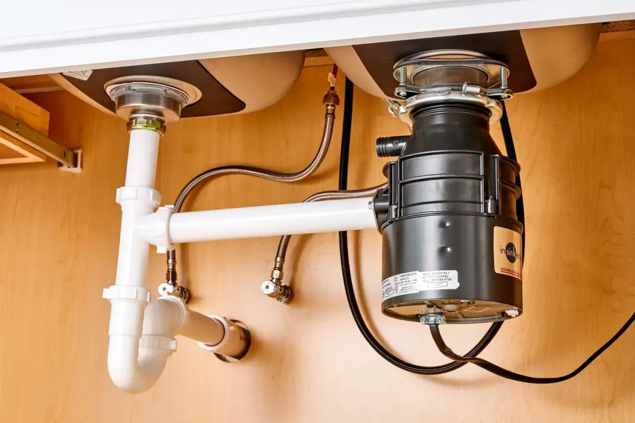 Under-sink view of a dual kitchen sink installation, showing plumbing pipes, a garbage disposal unit, and water supply lines.