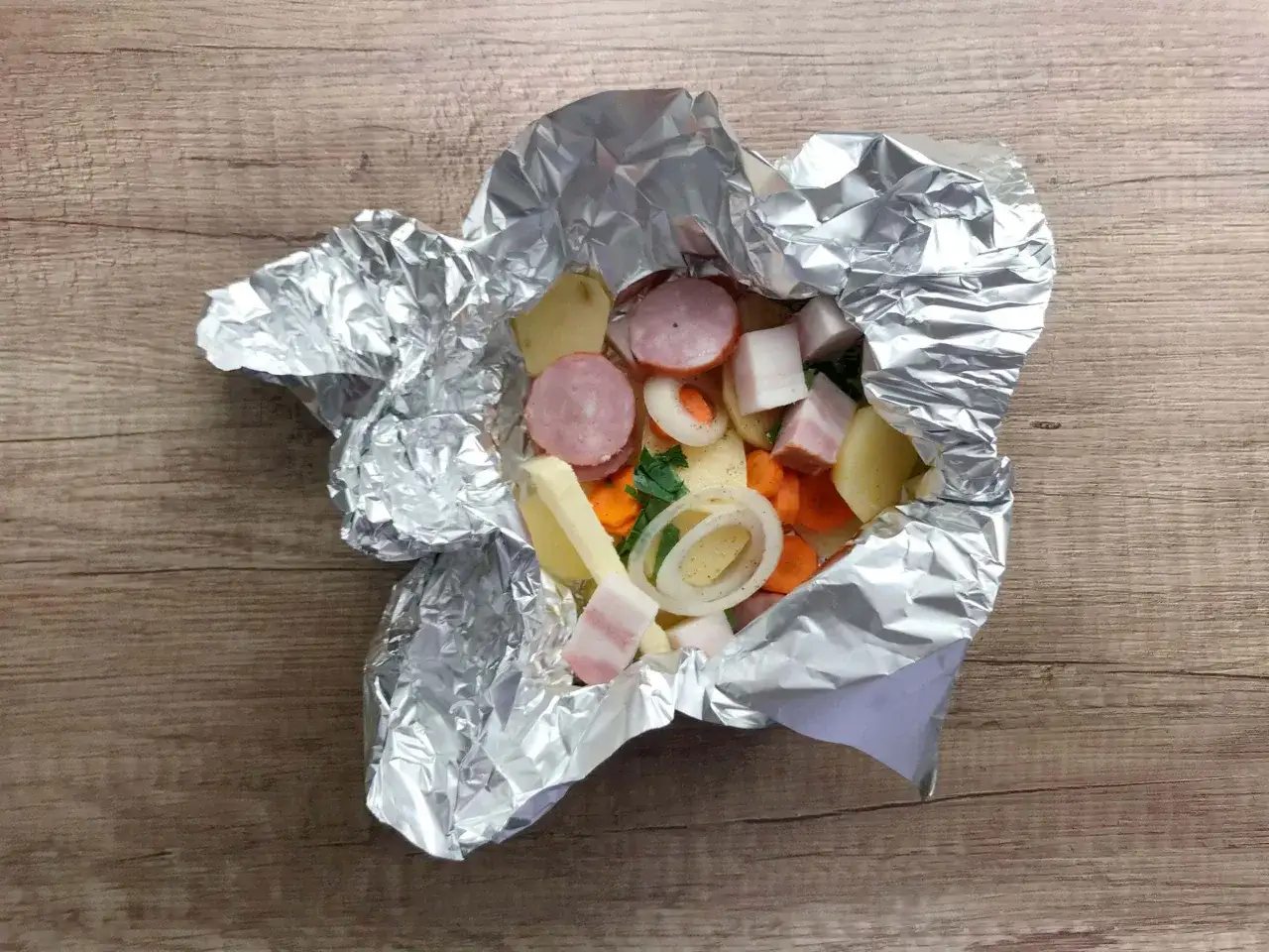 Folia aluminiowa z gotowymi do grillowania ziemniakami, kiełbasą, boczkiem i warzywami.