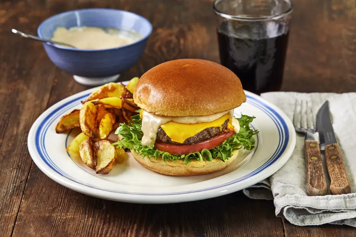 En saftig hamburgare med ost, sallad och tomat, serverad med klyftpotatis och en skål med hemlagad hamburgerdressing.