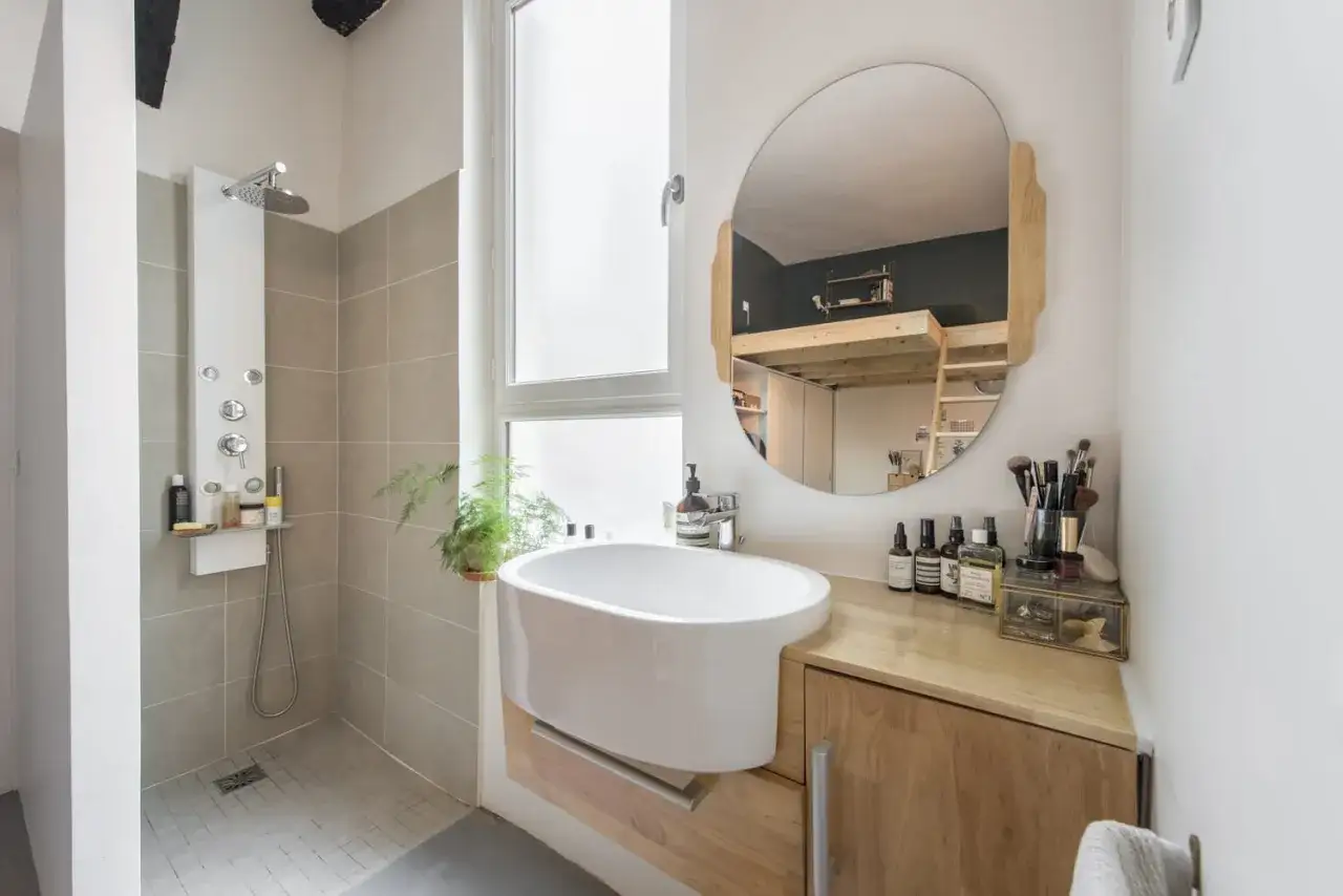 Salle de bain blanc et bois, avec douche moderne, vasque design et miroir rond refl&eacute;tant un lit mezzanine.
