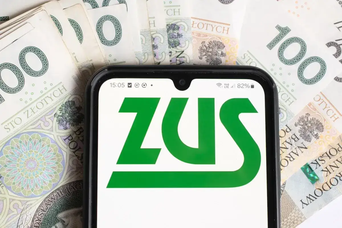 Sprawdzasz, ile wynosi renta socjalna? Na ekranie telefonu logo ZUS na tle polskich stu-złotówek.