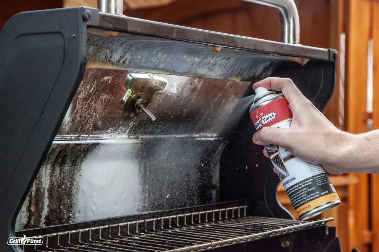Hand spr&uuml;ht Reinigungsschaum auf einen schmutzigen Weber Gasgrill, um ihn zu reinigen.