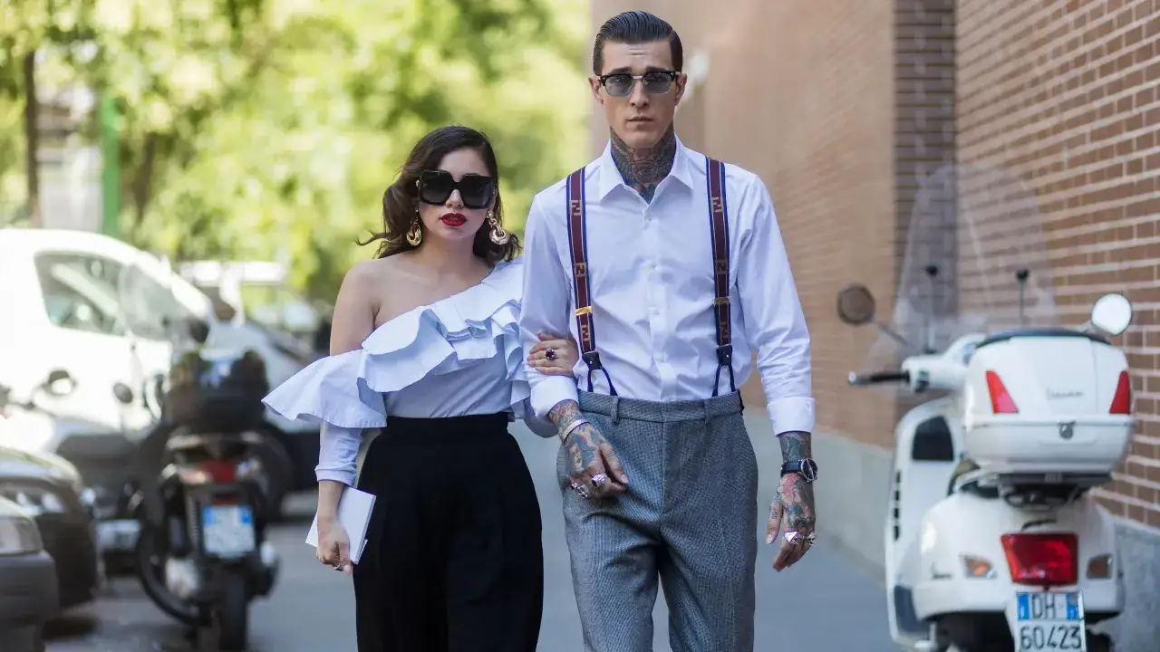 Un invitado boda hombre sin traje, con tirantes y tatuajes, camina junto a una mujer con un voluminoso top blanco y pantalones negros.