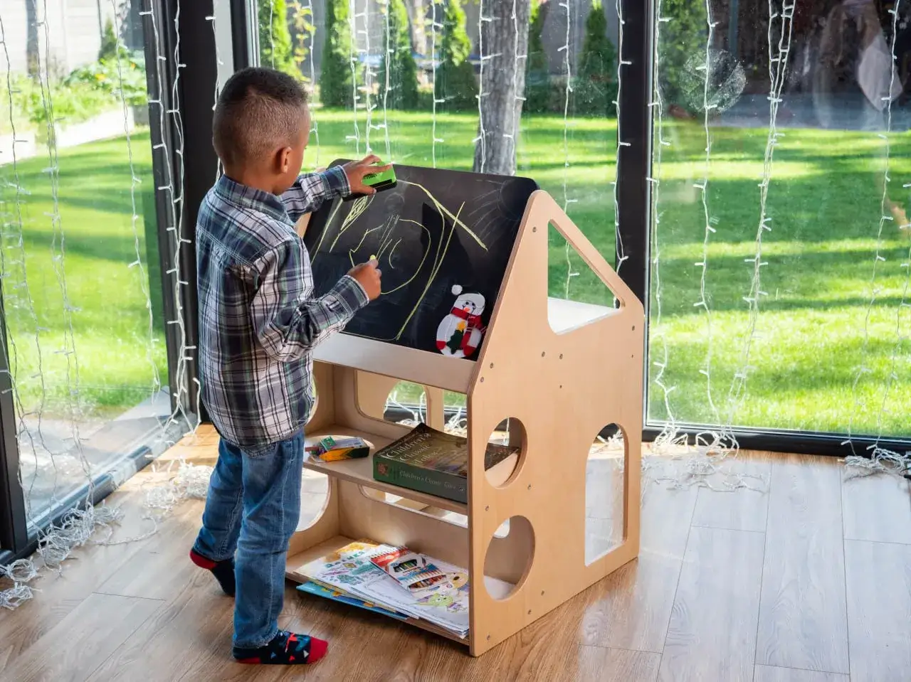 A boy draws on a chalkboard easel, part of a Montessori backyard playhouse. Books and art supplies are neatly stored below.