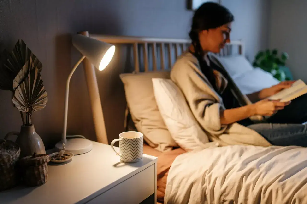 Frau liest im Bett bei Nachttischlampe, eine Tasse Tee daneben. Ein gemütlicher Abend mit Hausmitteln für besser schlafen.
