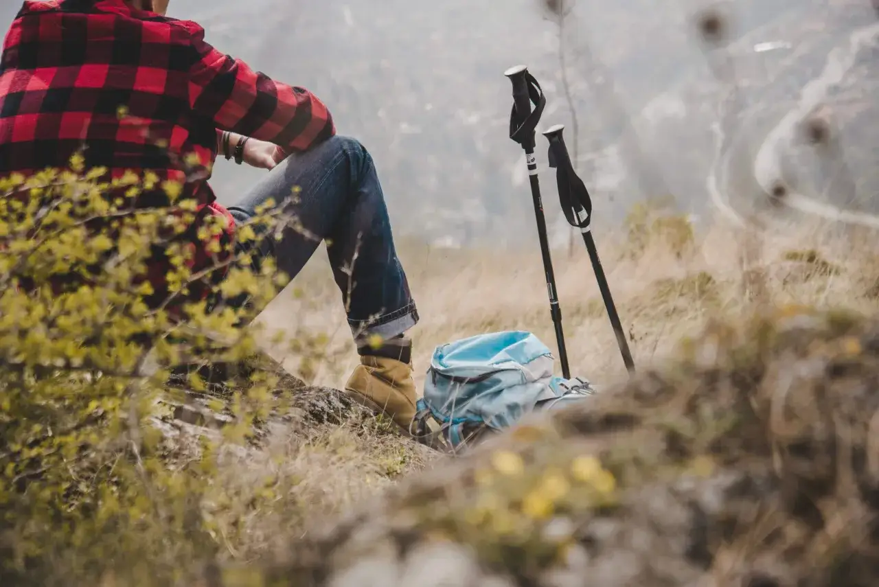 Osoba w kratę siedzi na skale, obok plecak i kije trekkingowe. To inspiracja, jak spakować się na wędrówkę po górach opawskich lista kontrolna.