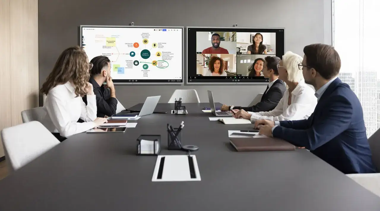 A team uses board management software for a hybrid meeting, with some in person and others on a video call, discussing ideas on a large screen.
