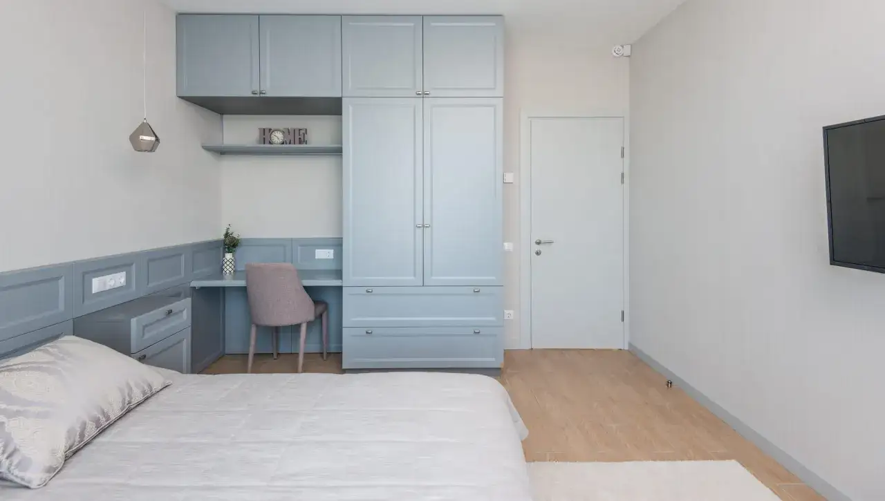 Chambre d'amis polyvalente avec lit, bureau int&eacute;gr&eacute;, armoire bleue et t&eacute;l&eacute;vision murale.