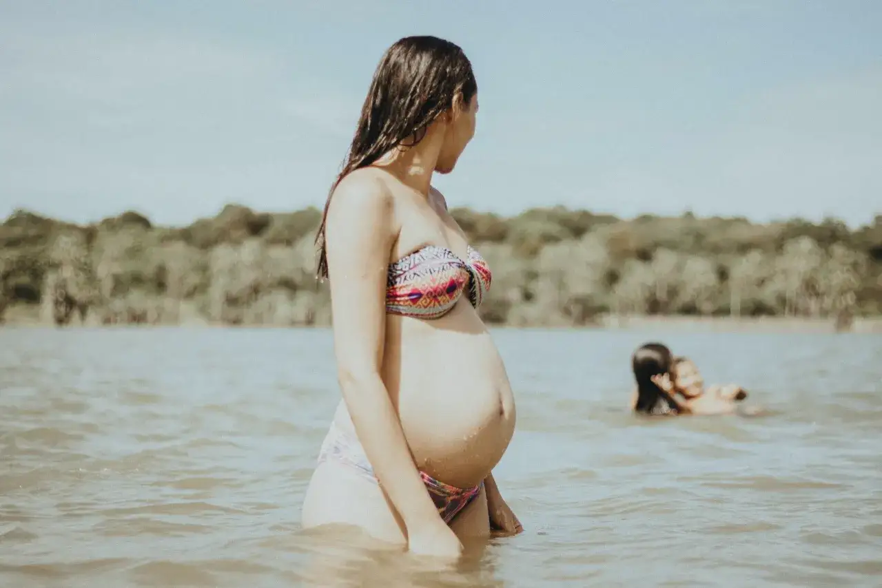 En gravid kvinna i en f&auml;rgglad bikini st&aring;r i vattnet, med en skogskl&auml;dd strand i bakgrunden. Tv&aring; barn leker i vattnet l&auml;ngre bort.