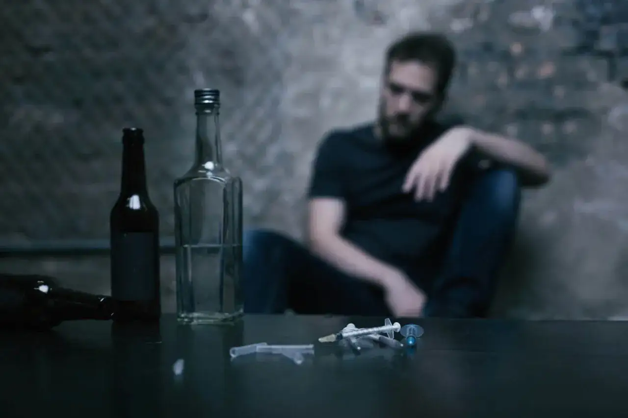 Mężczyzna siedzi w kącie, obok butelki i strzykawki. Jego wzrok jest pusty, jakby był suchym alkoholikiem, który stracił nadzieję.