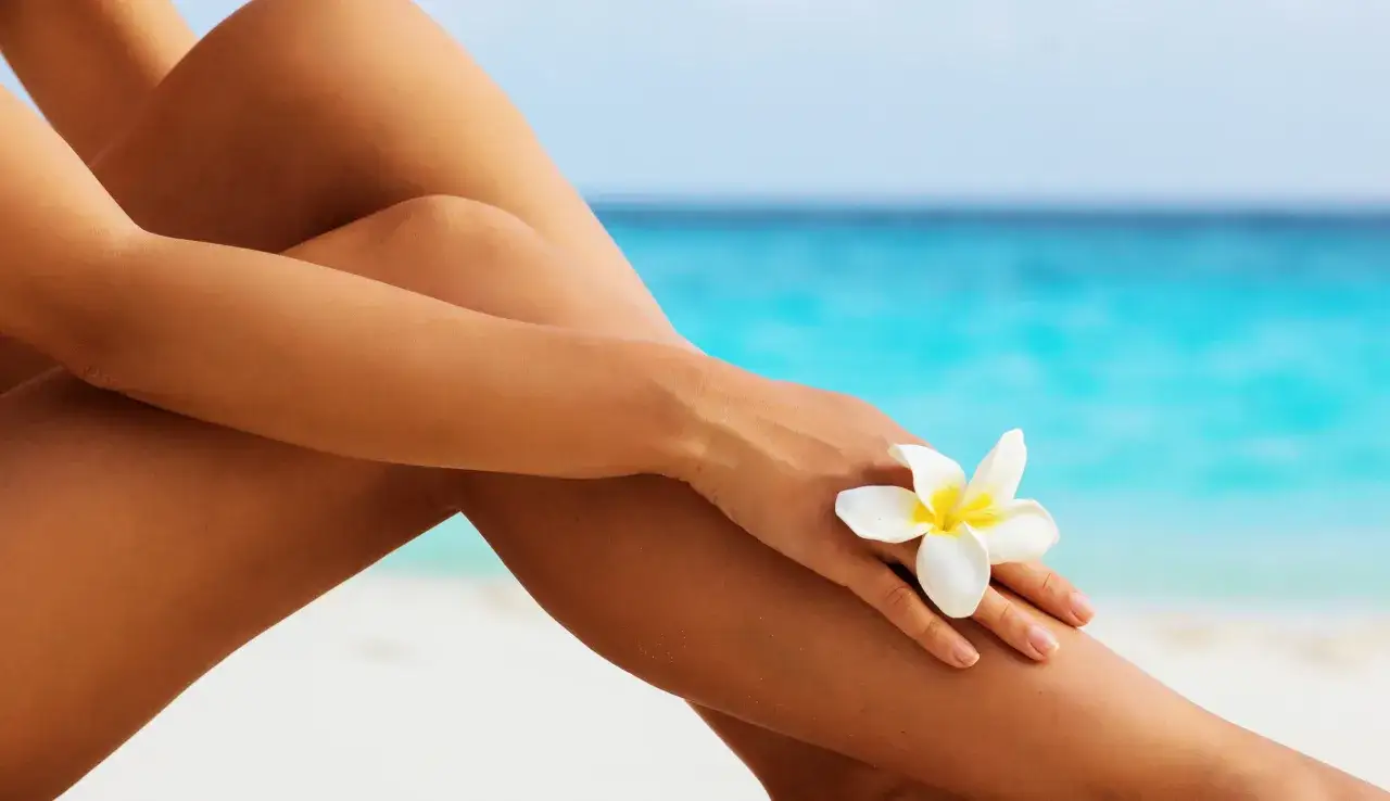 Gambe abbronzate al sole, con un fiore bianco appoggiato sulla pelle. Il mare turchese sullo sfondo evoca il desiderio di come abbronzarsi in vacanza.