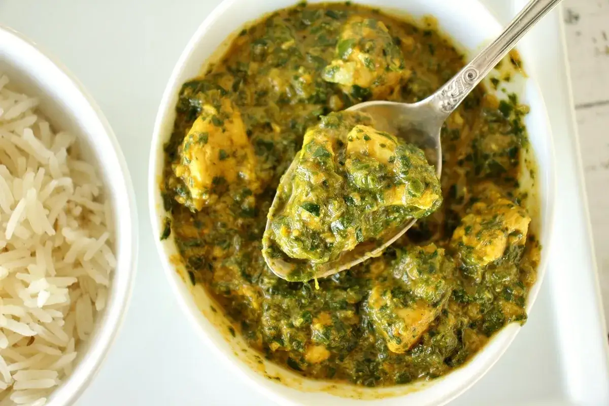En sked lyfter upp krämig palak chicken, en smakrik kycklingrätt med spenat, bredvid en skål med fluffigt ris.
