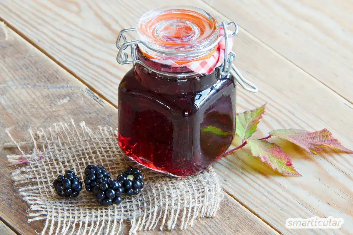 Glas mit dunklem Brombeergelee, daneben frische Brombeeren und Blätter. Perfekt, um das Brombeergelee zu verfeinern.