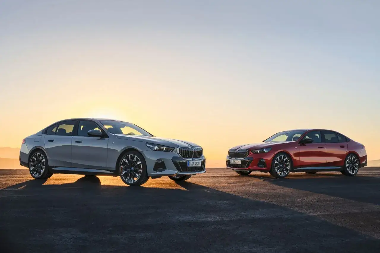 Dwa nowe BMW G60, srebrne i czerwone, stoją na tle zachodzącego słońca.