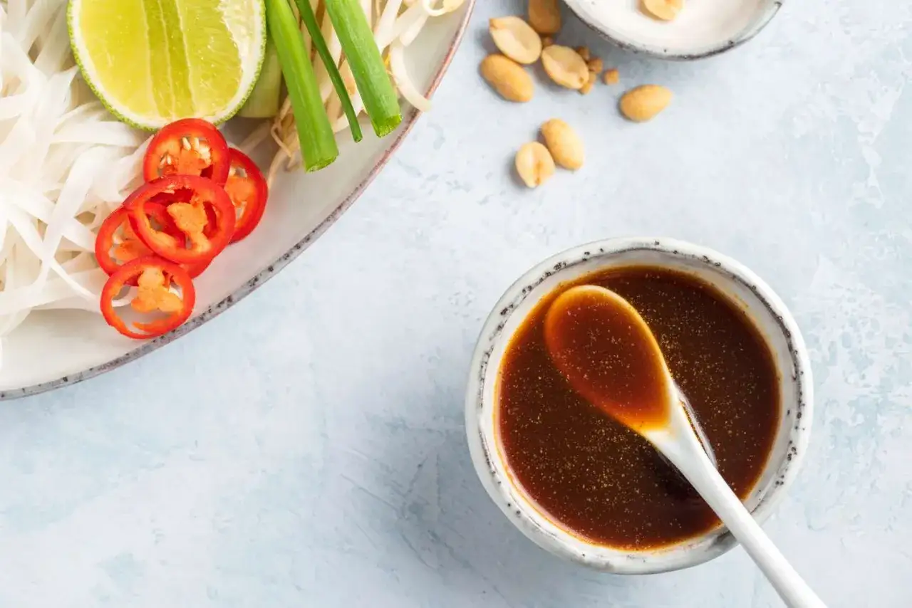 Miseczka z aromatycznym **pad thai sosem**, obok talerz z makaronem ryżowym, limonką, chili i szczypiorkiem.