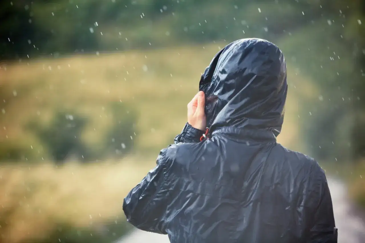 Person im Regen, die Kapuze einer Jacke mit PU-Beschichtung festh&auml;lt.