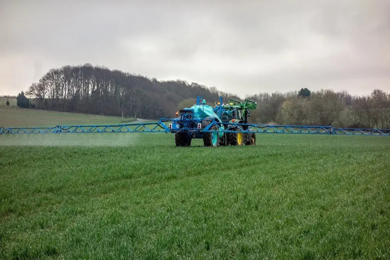 Traktor z opryskiwaczem, prawdopodobnie z preparatem **lentipur**, pracuje na zielonym polu. W tle widać drzewa i wzgórza.