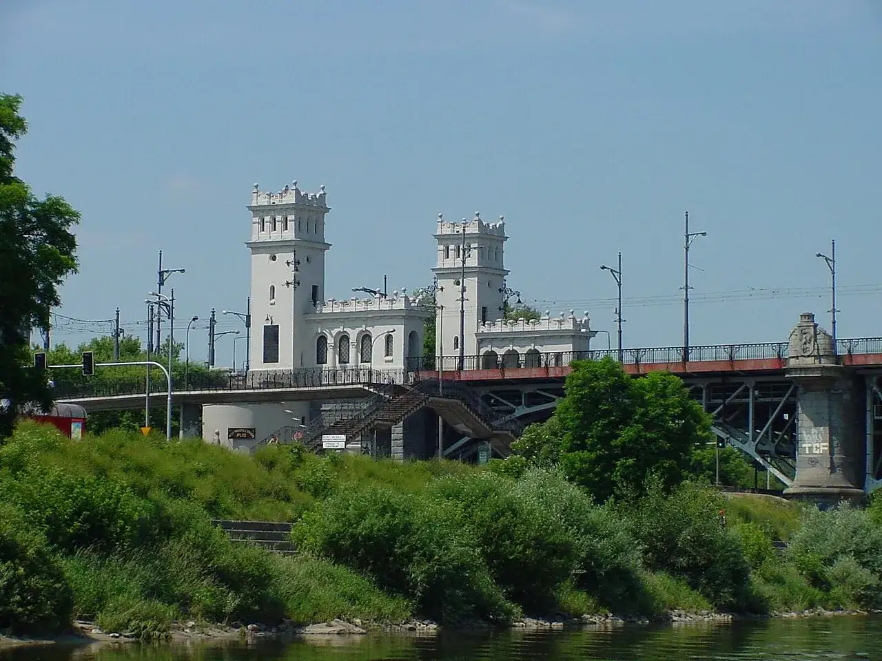 Białe wieże i arkadowe budowle nad Wisłą, część Mostu Poniatowskiego, z zielonymi skarpami i wodą w tle.