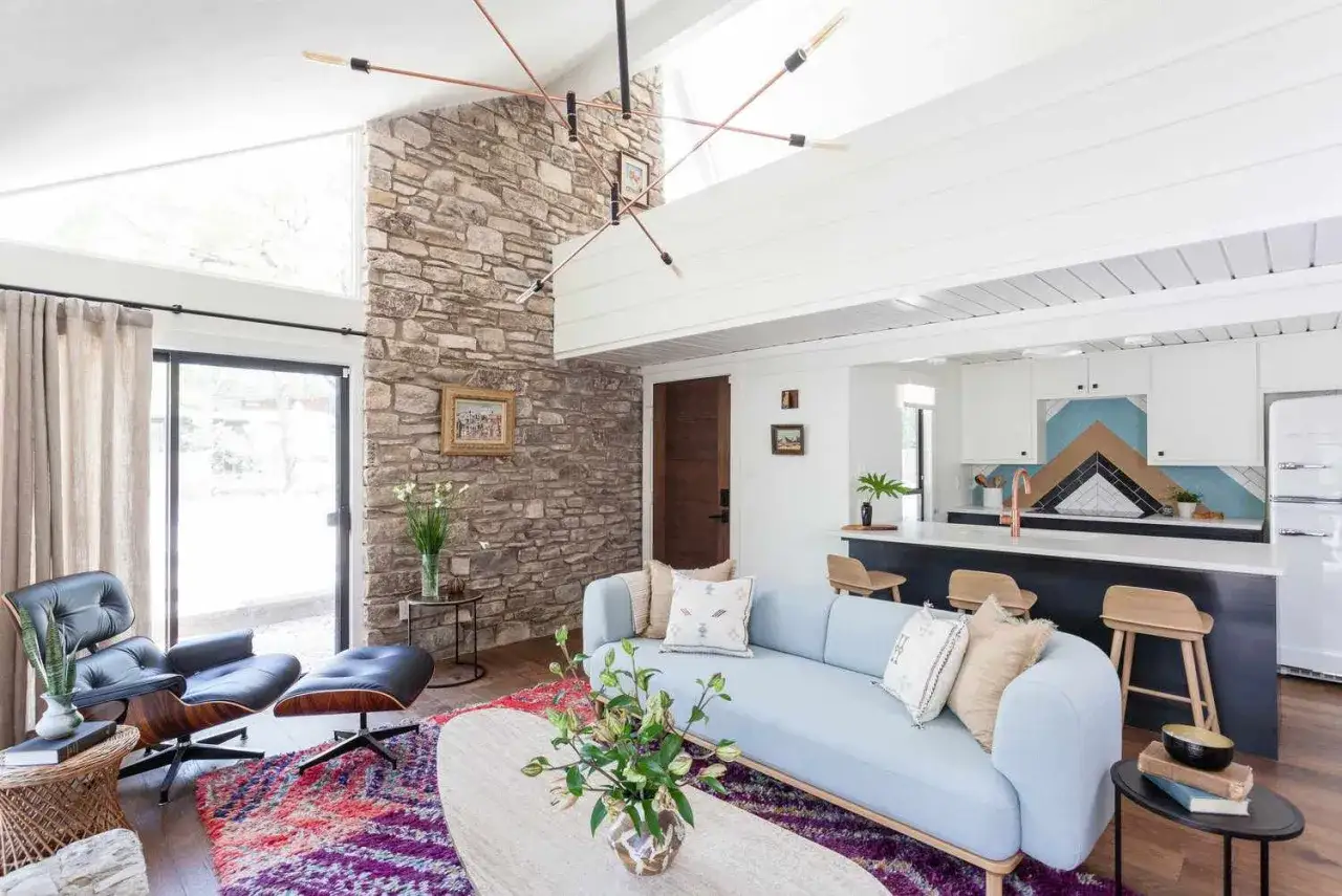Modernes wohnzimmer einrichten mit hellblauer Couch, Steinwand und bunter Teppich. Offene Küche mit Bar und Hockern.