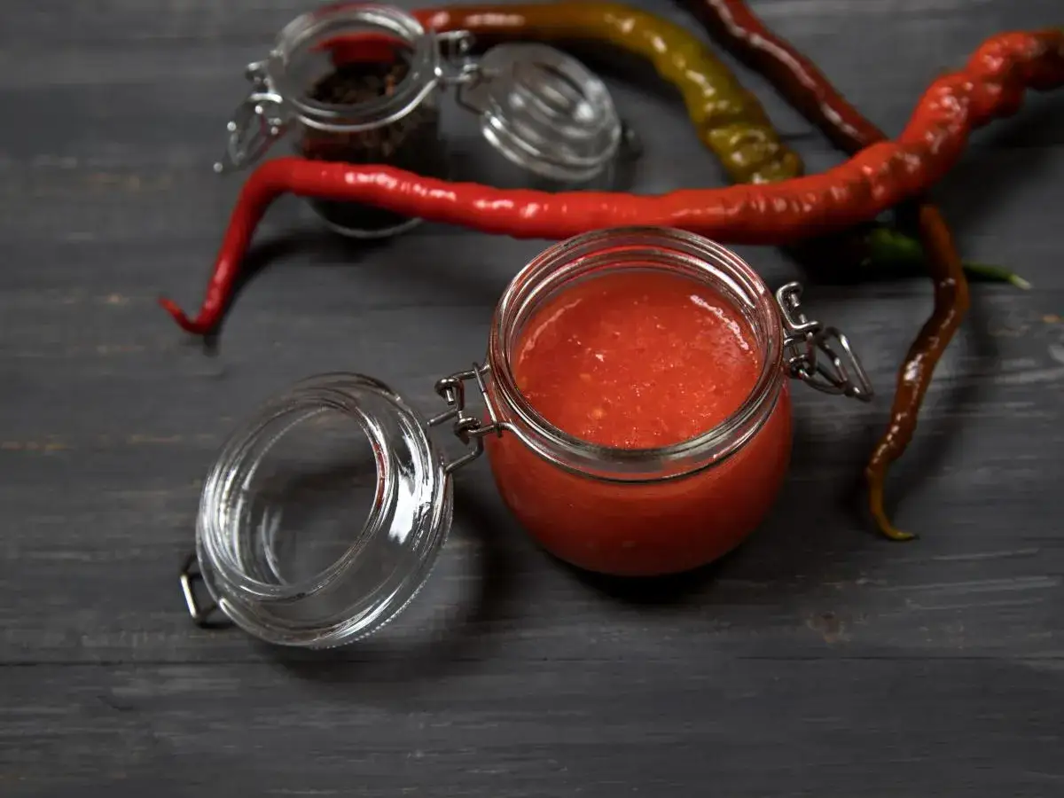 Domowy sos sriracha z papryczek chilli i czosnku w słoiku, otoczony świeżymi papryczkami i ziarnami pieprzu.