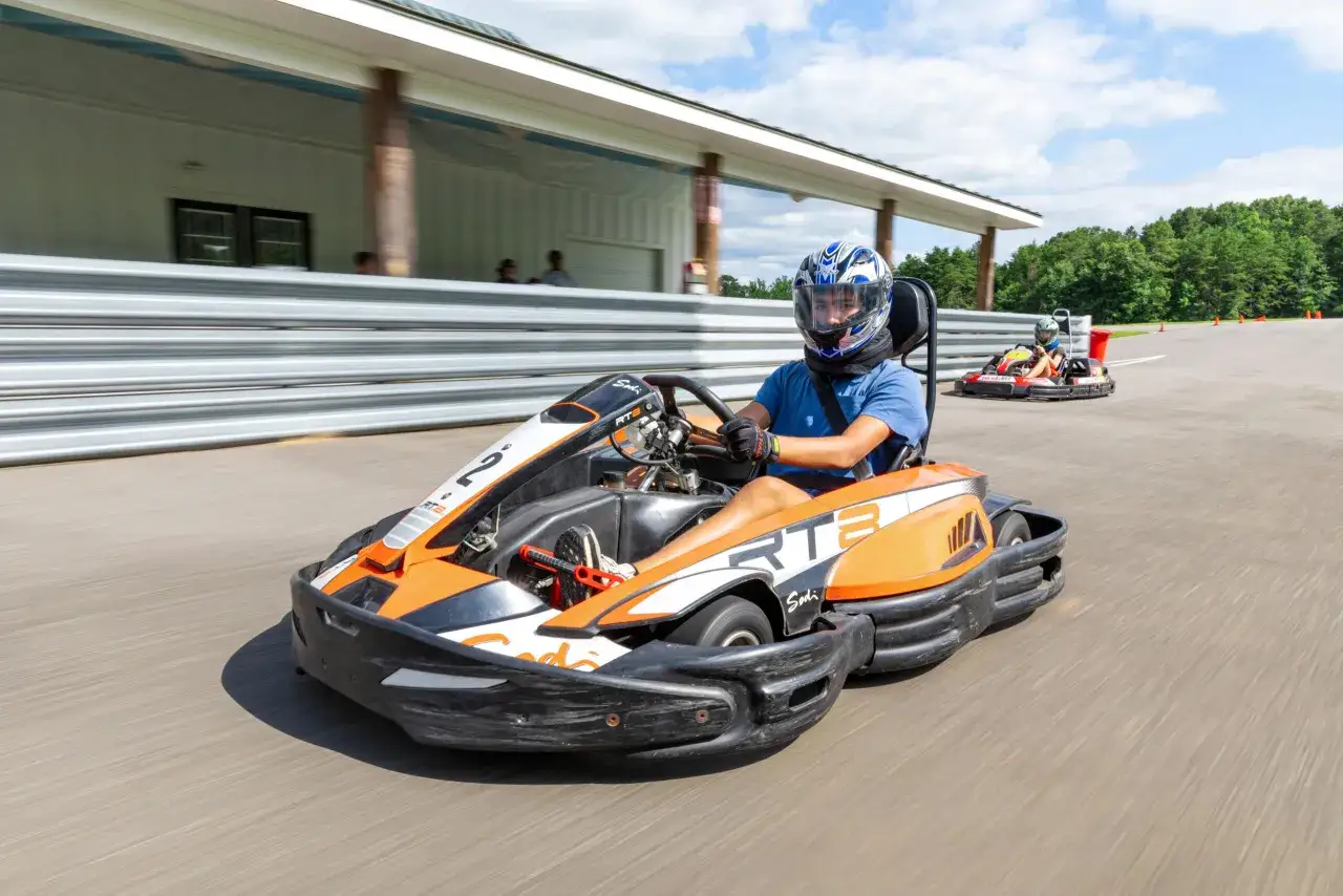 Un jeune homme en casque bleu et blanc pilote un kart orange et noir sur une piste de colo karting, suivi par un autre kart.