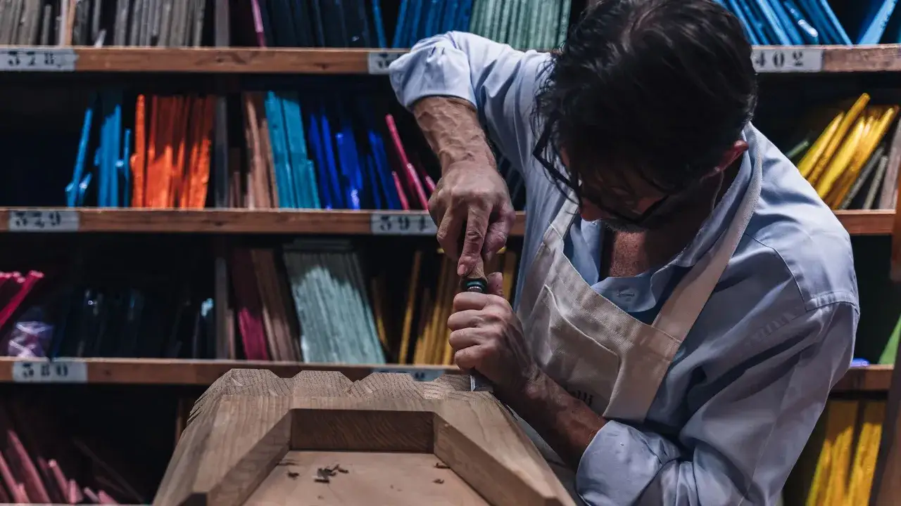 Artigiano al lavoro su un pezzo di legno, con scaffali pieni di campioni colorati alle spalle, un omaggio alla **fondazione Cologni**.