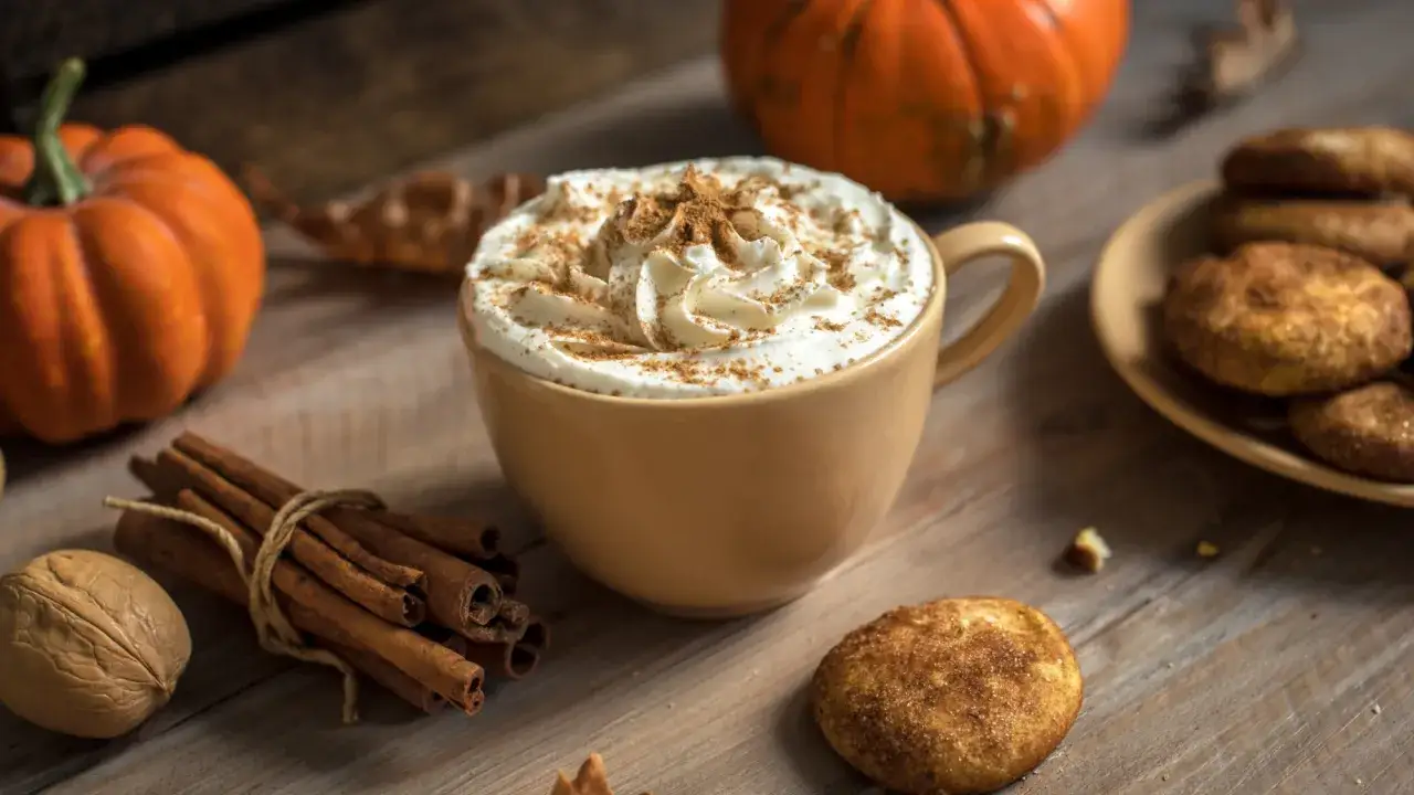 Jesienna kawa pumpkin spice latte z bitą śmietaną i cynamonem, obok dynie i ciasteczka.
