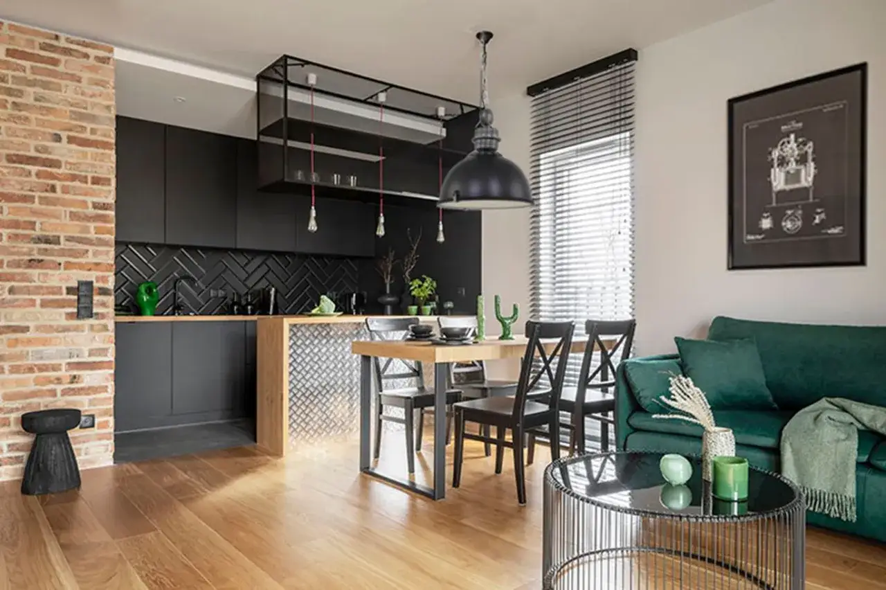 Nowoczesna kuchnia w stylu loft z czarnymi frontami, ceglaną ścianą i drewnianym blatem. Obok jadalnia z drewnianym stołem i krzesłami.