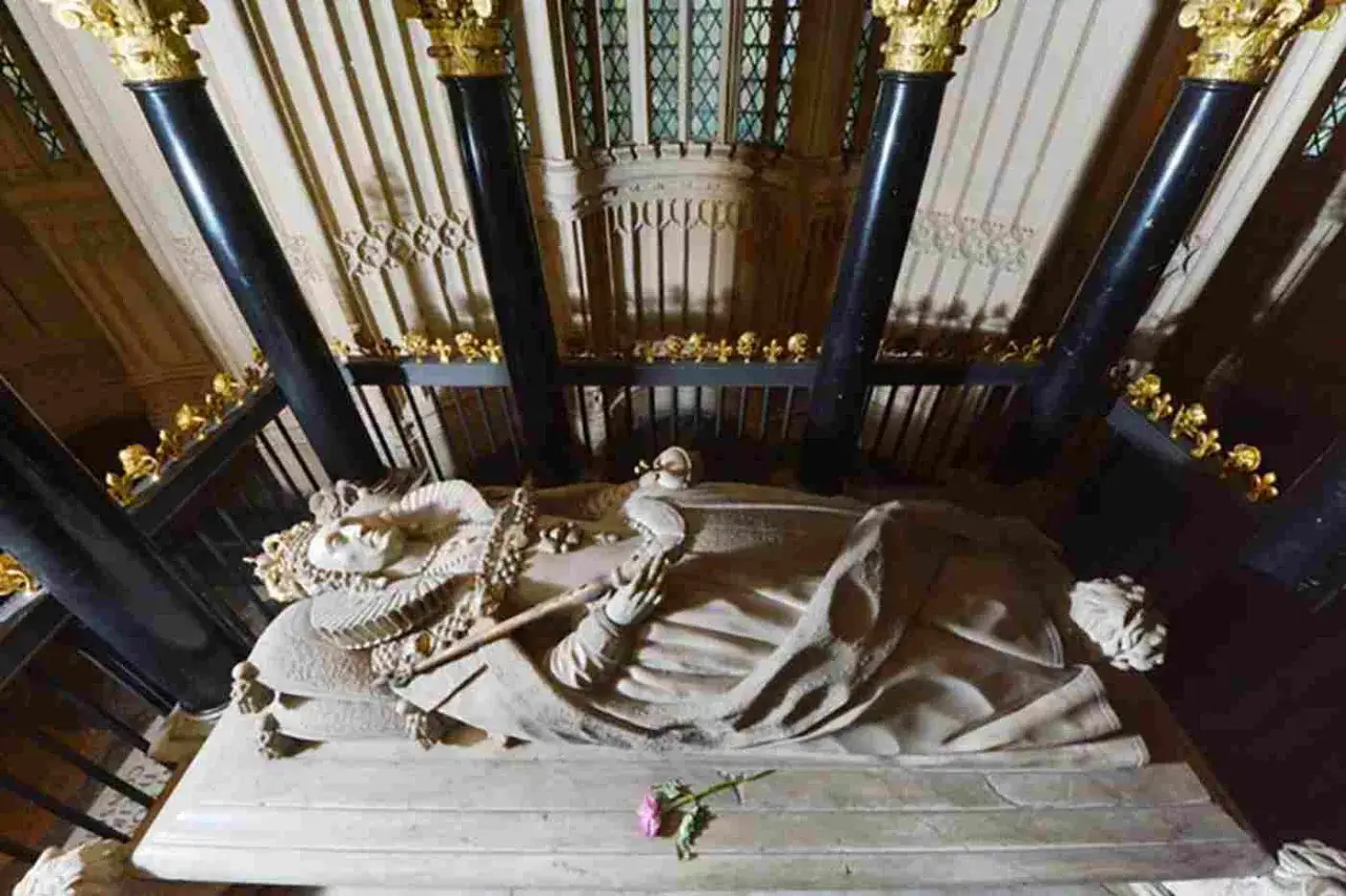 A stone effigy of a queen lies in state, a testament to the grandeur of abbey graves. A single pink rose rests at her feet.