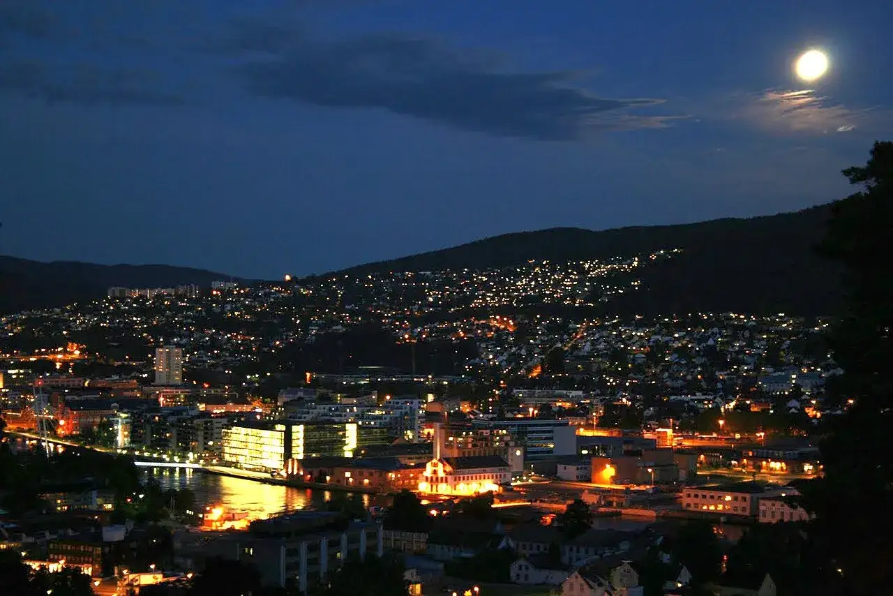 Nocny widok na Drammen w Norwegii, z rozświetlonymi domami na wzg&oacute;rzach i księżycem na niebie.