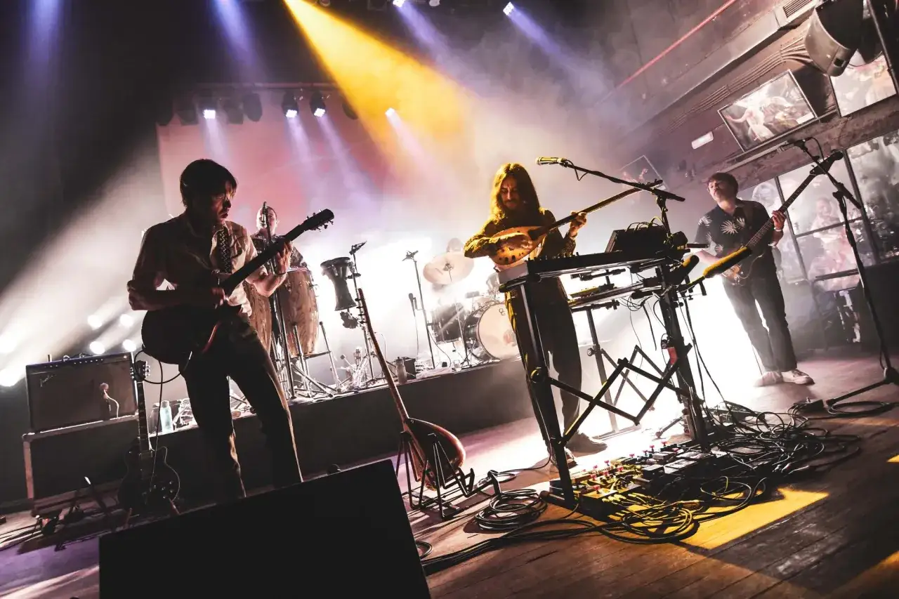 Banda tocando en vivo, con guitarras, teclados y percusión. La iluminación crea una atmósfera de concierto, como si los **track dogs** estuvieran persiguiendo el ritmo.