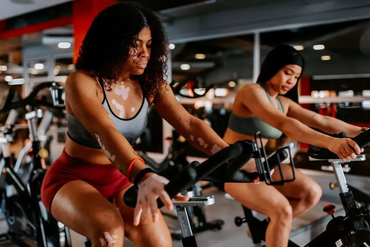 Two women engage in high intensity cycling in a gym, pushing their limits on stationary bikes.