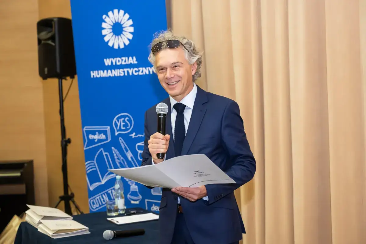 Mężczyzna w garniturze przemawia na tle banera Wydziału Humanistycznego, inspirując do wyboru studiów po humanie.