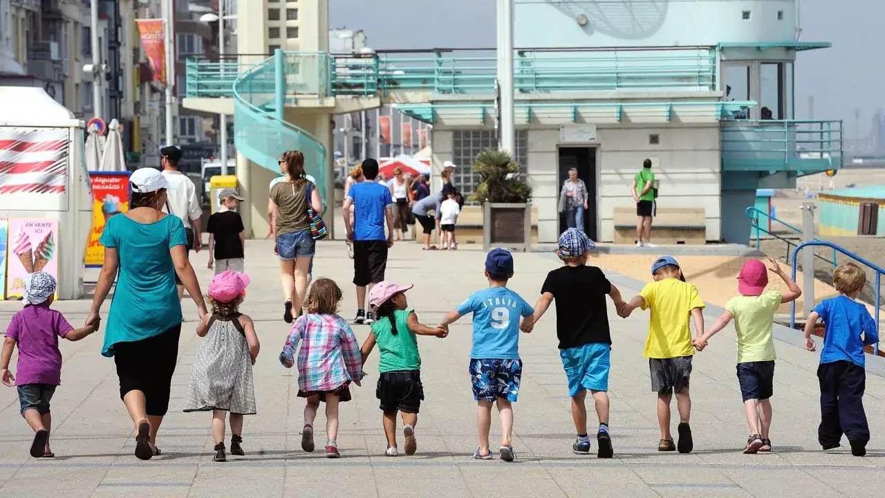 Un groupe d'enfants se promène main dans la main, profitant d'une journée ensoleillée. L'encadrement des enfants est assuré par des adultes attentifs, créant une scène de joie et de sécurité.