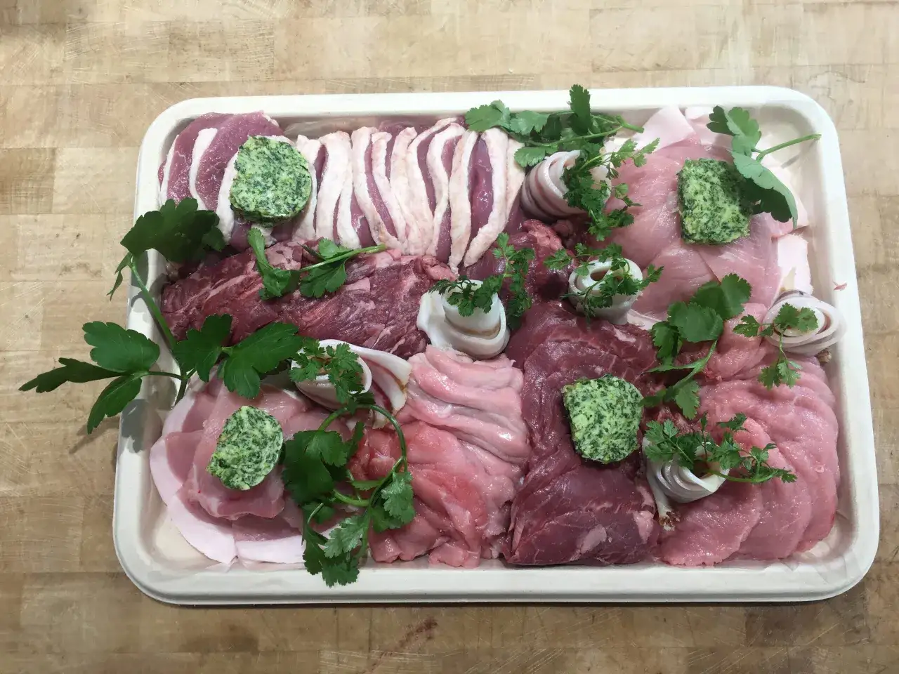 Plateau de pierrade pour 10 personnes : assortiment de viandes crues, de beurre persillé et de persil frais, prêt à être cuisiné.