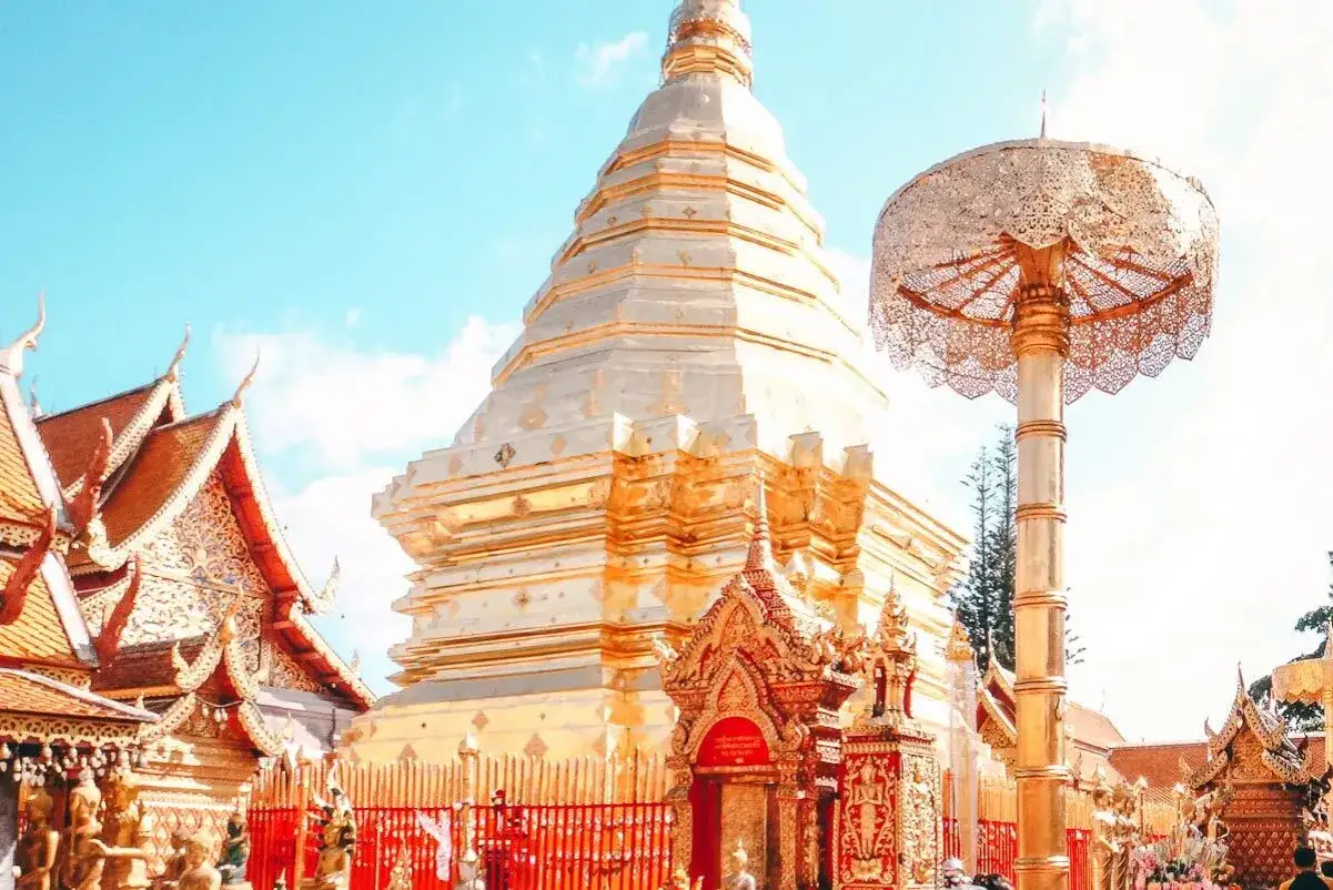 Goldene Stupas und Tempeld&auml;cher in Chiang Mai unter blauem Himmel. Ein filigraner Sonnenschirm steht daneben.