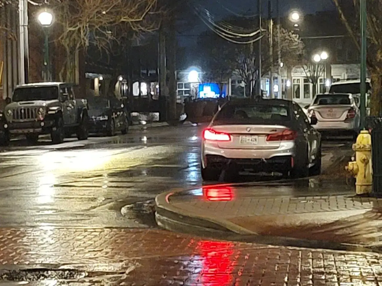 Srebrny sedan z włączonymi światłami postojowymi stoi na mokrej ulicy. Obok niego Jeep i inne samochody.