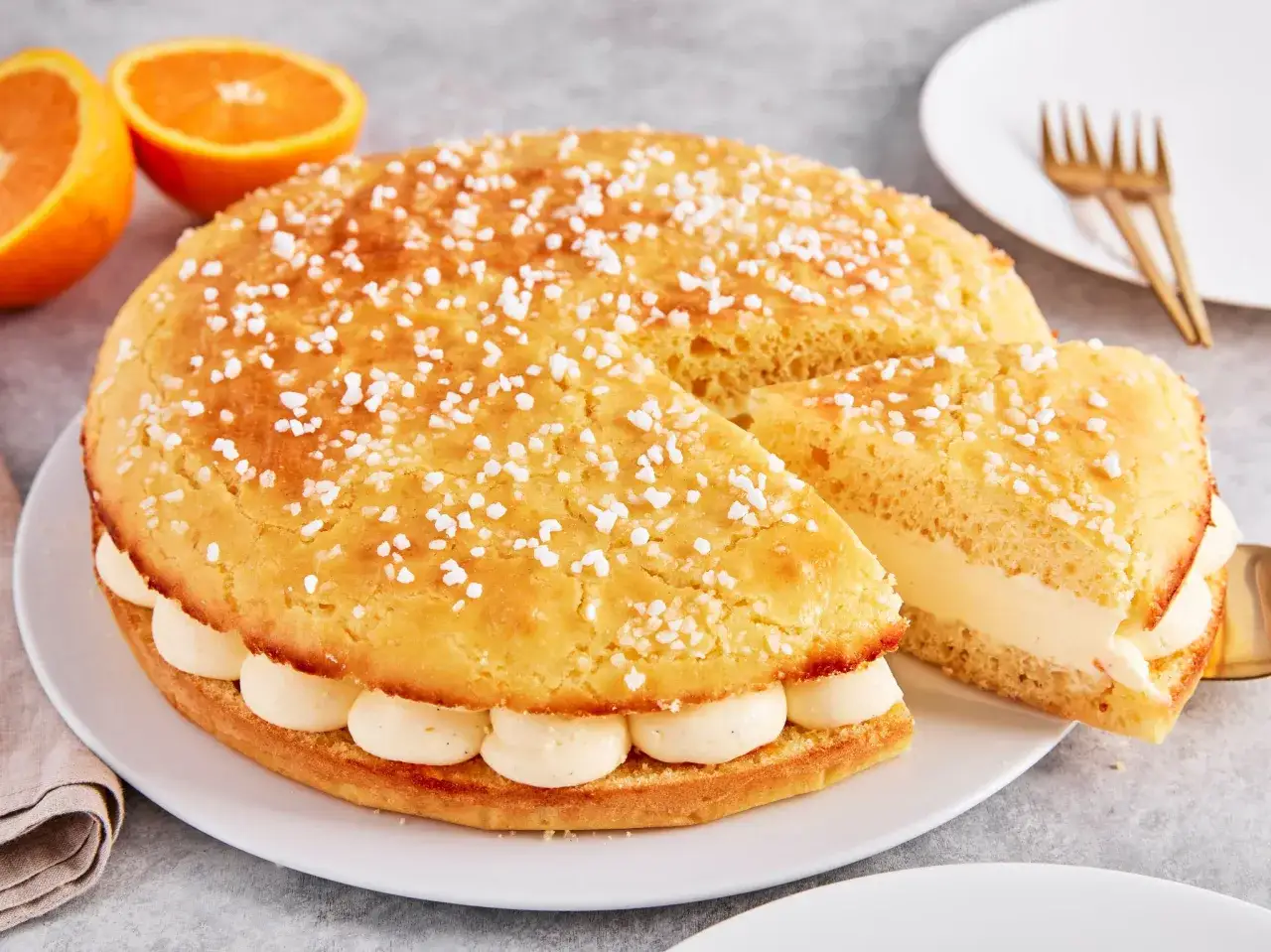 Eine köstliche Tarte Tropezienne, bestreut mit Zuckerkristallen, gefüllt mit cremiger Vanillecreme. Orangen liegen daneben.