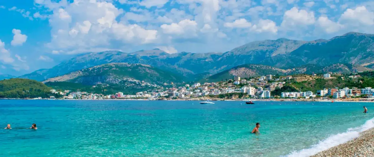 Albania - wczasy samolotem all inclusive. Turkusowe morze, plaża, miasteczko na wzgórzu i góry w tle. Idealne miejsce na relaks.