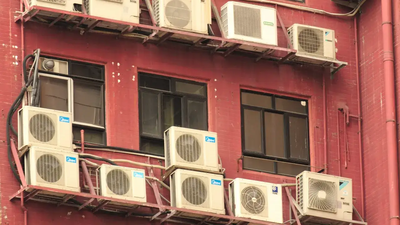 Wiele klimatyzatorów zamontowanych na czerwonej ścianie budynku. Czy balkon jest częścią wspólną, trudno stwierdzić po tych jednostkach.