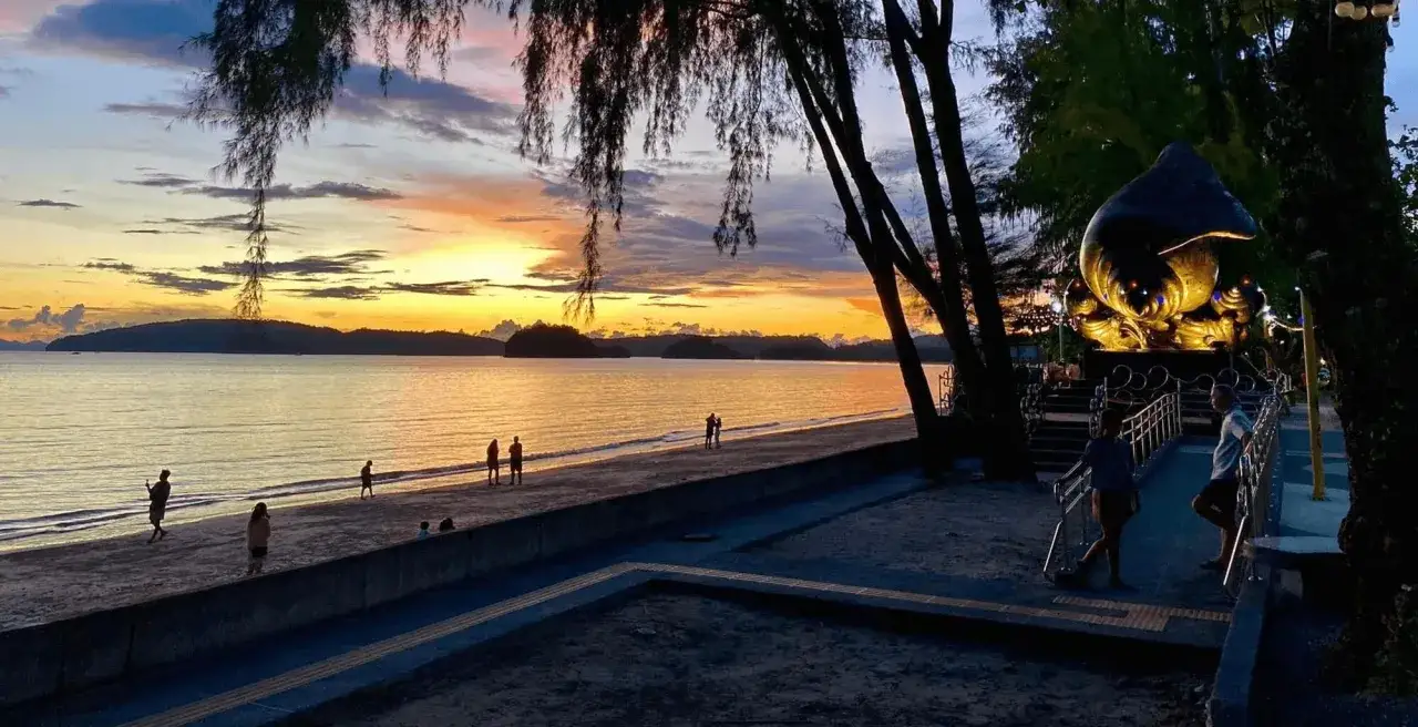 Zachód słońca nad Ao Nang Beach. Ludzie spacerują po piasku, a w oddali widać rzeźbę.