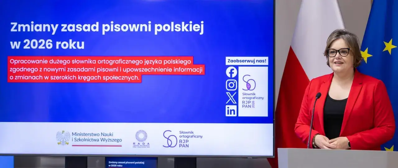 Kobieta w czerwonej marynarce przemawia na tle ekranu z napisem 