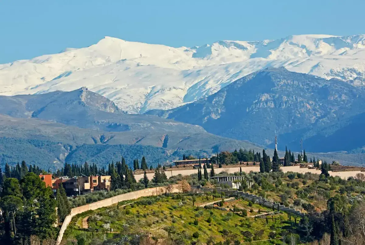 Widok na ośnieżone góry Sierra Nevada, gdzie można jeździć na nartach w Hiszpanii. Na pierwszym planie zielone wzgórza z budynkami i cyprysami.