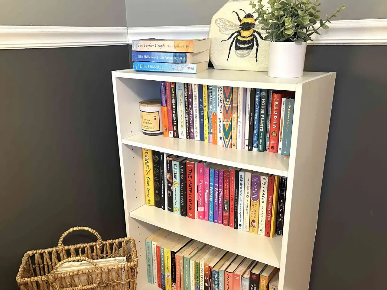 Une &eacute;tag&egrave;re blanche remplie de livres de poche, parfaite pour fabriquer une biblioth&egrave;que pour livres de poche. Un panier en osier et une plante verte compl&egrave;tent la sc&egrave;ne.