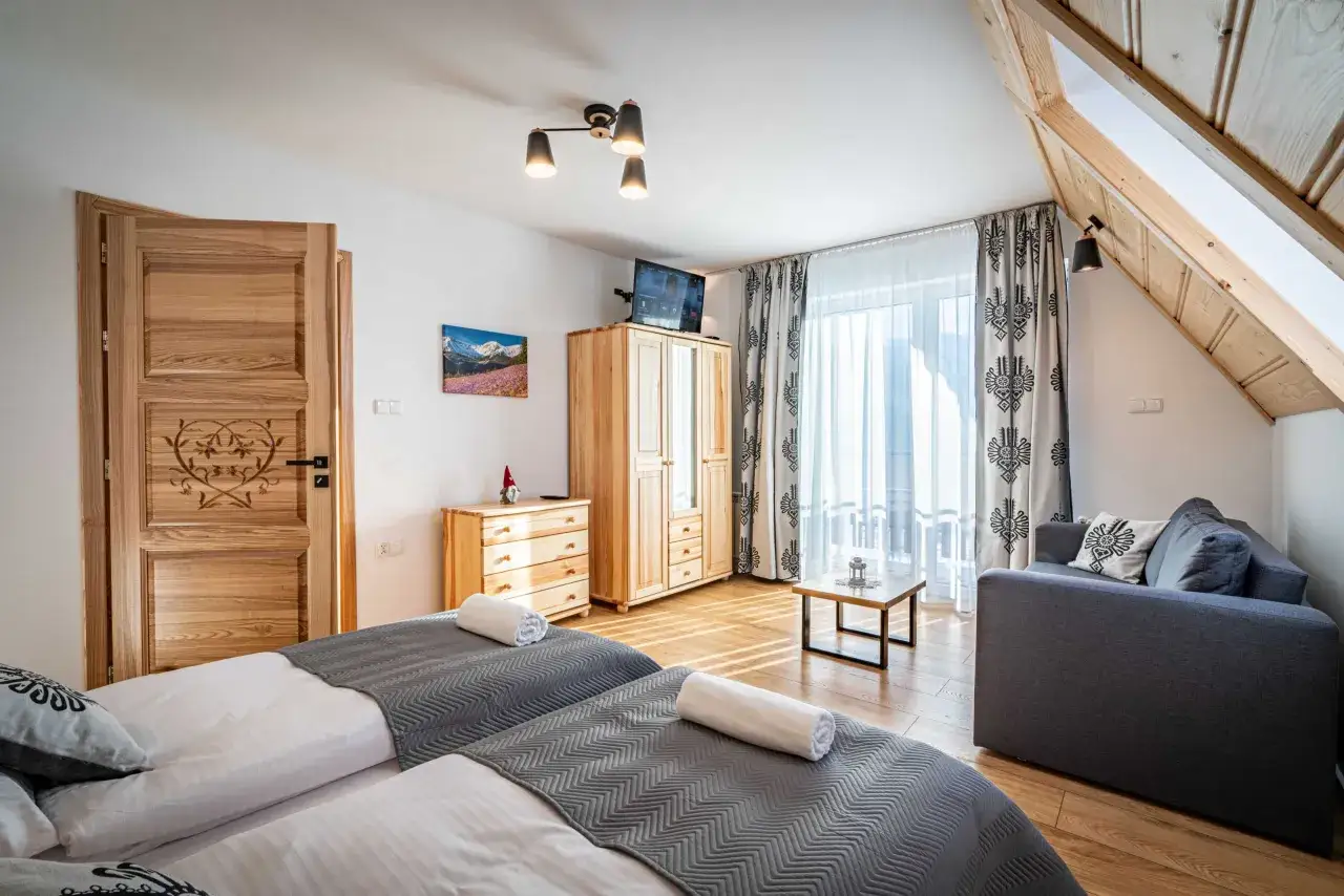Przytulny apartament pod wierchem. Dwa łóżka, kanapa, drewniane meble i góralski akcent na drzwiach.