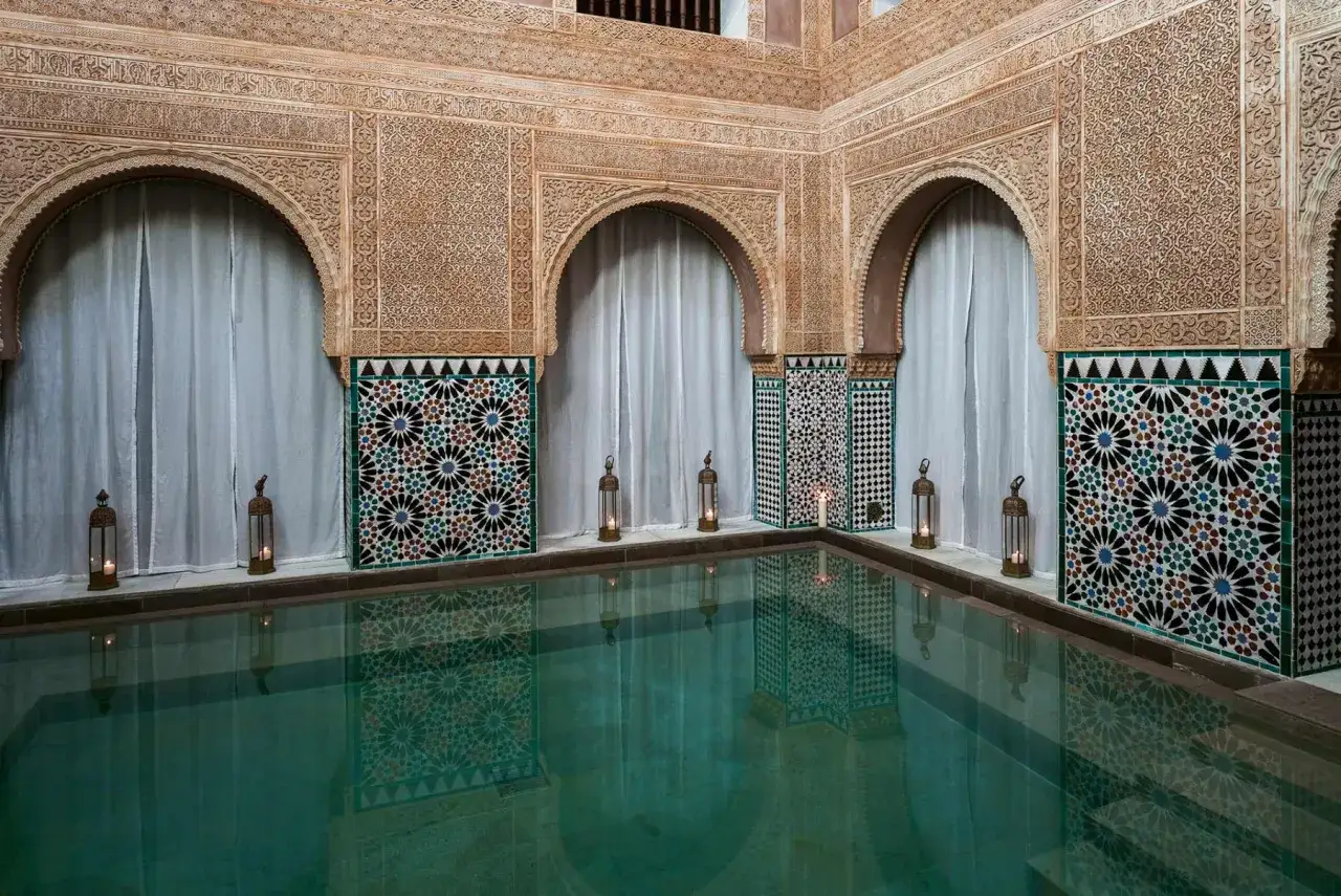 Ein **Hammam Al Andalus** mit t&uuml;rkisfarbenem Wasser, kunstvollen Mosaiken und filigranen Stuckarbeiten. Kerzenlicht in Laternen taucht den Raum in warmes Licht.
