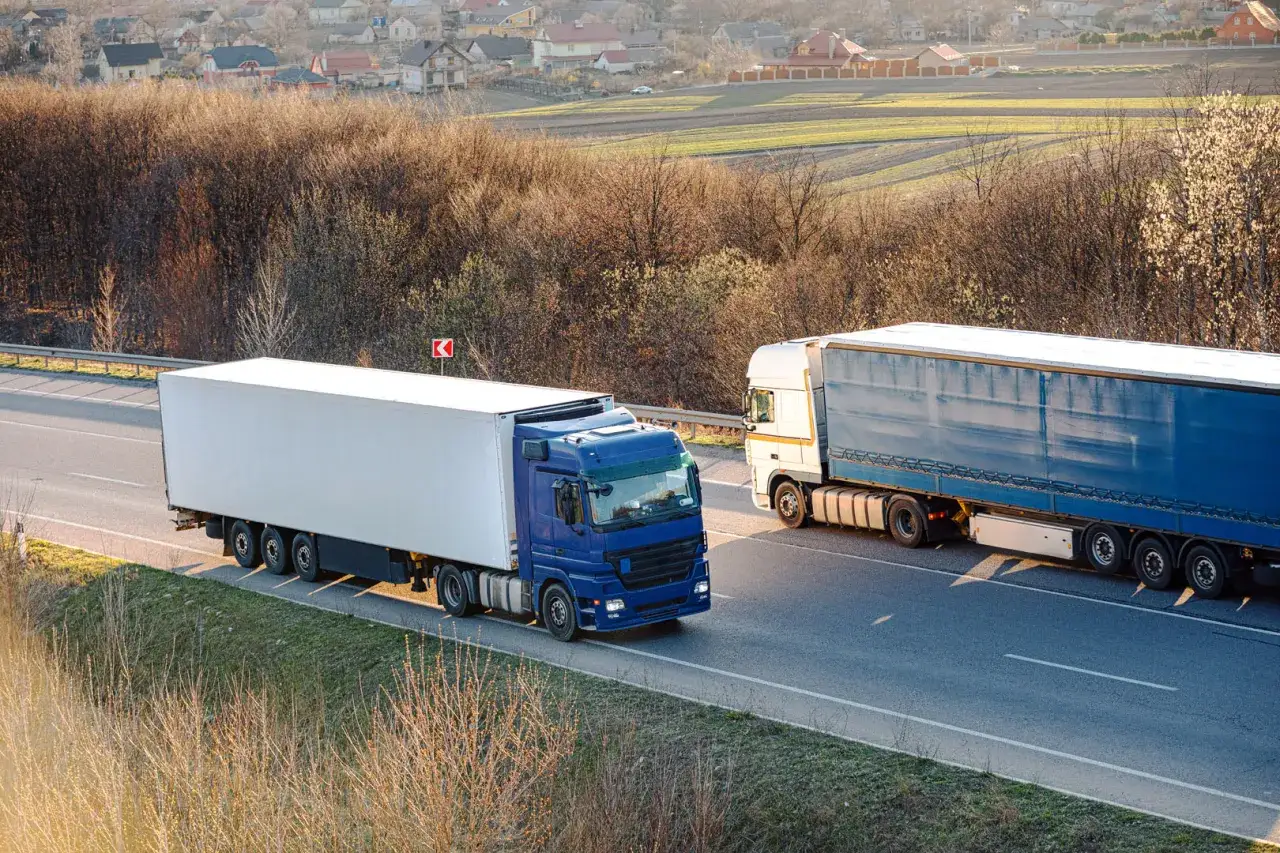 Dwa ciężkie samochody ciężarowe, jeden niebieski, drugi biały, jadą autostradą. To obraz transportu i spedycji w ruchu.