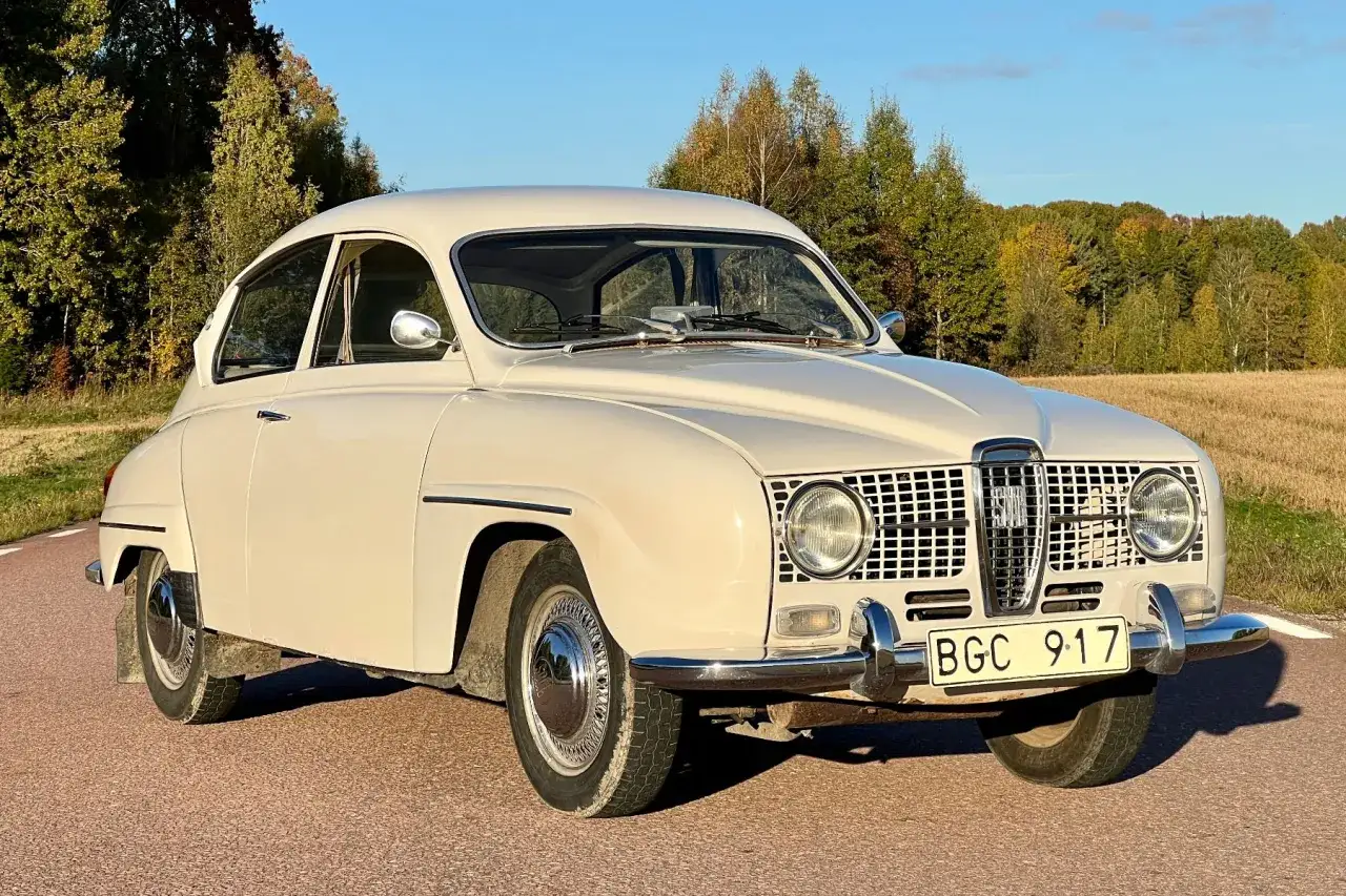 En krämvit Saab 2-takt står parkerad vid en vägkant med höstfärgad skog i bakgrunden.