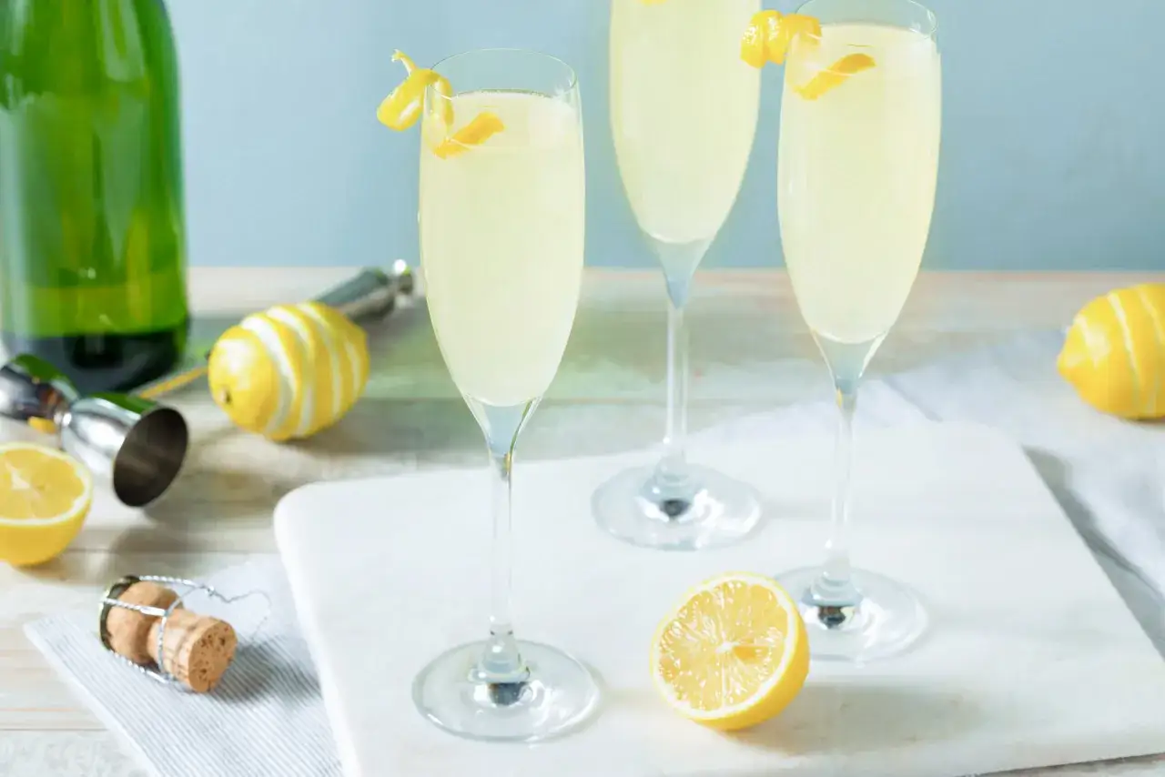 Drei elegante Gläser mit einem erfrischenden Champagner Cocktail, garniert mit Zitronenzesten. Daneben liegen frische Zitronen und ein Korken.