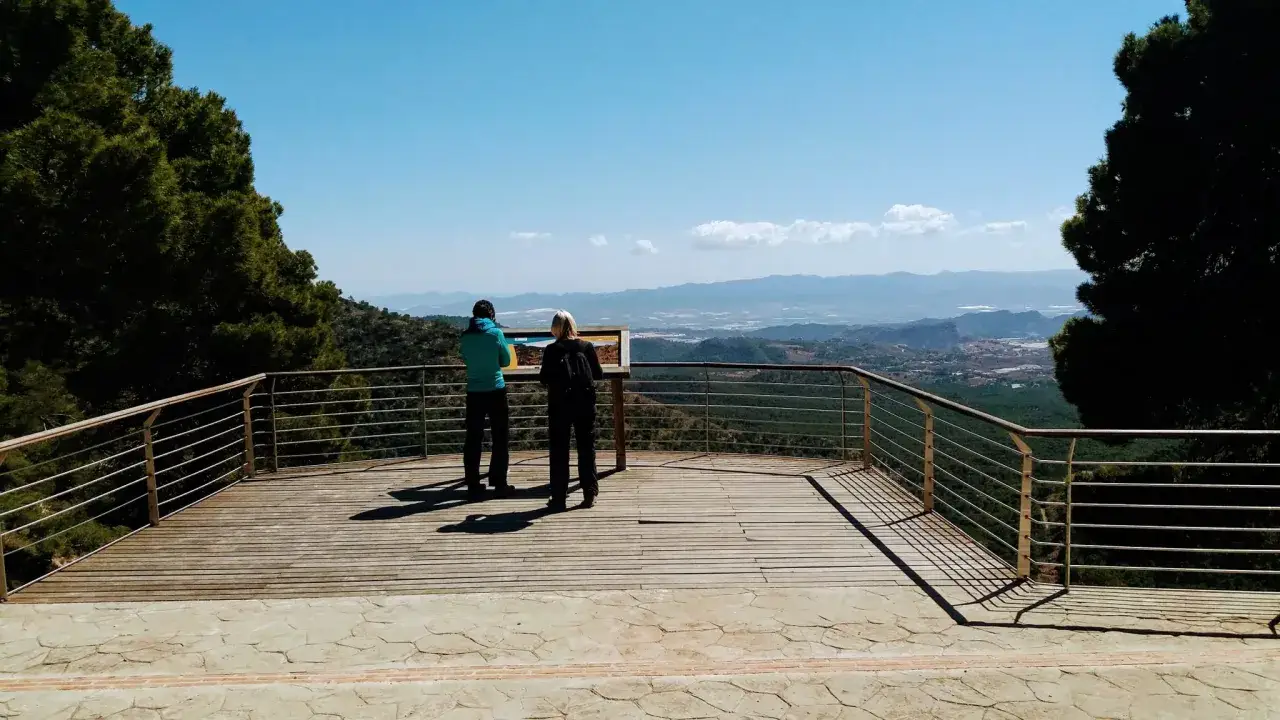 Dos personas observan un panel informativo en un mirador con vistas al **estrecho de la Arboleja** y un valle verde.
